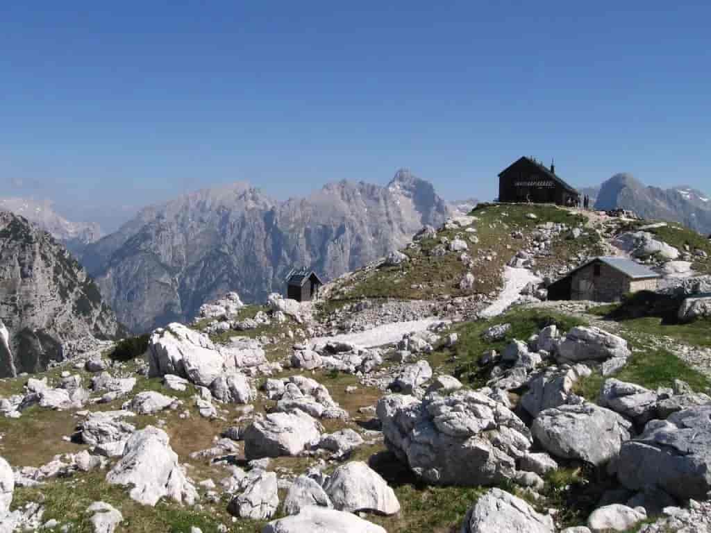 Zasavska Hut at Prehodavci on rocky alpine terrain with distant mountain range under clear blue sky.