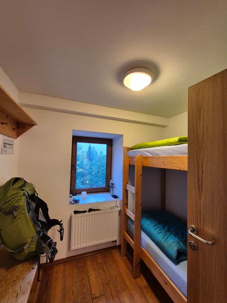 Bunk room interior with wooden floors, a large green backpack, and a window showing a forest outside.