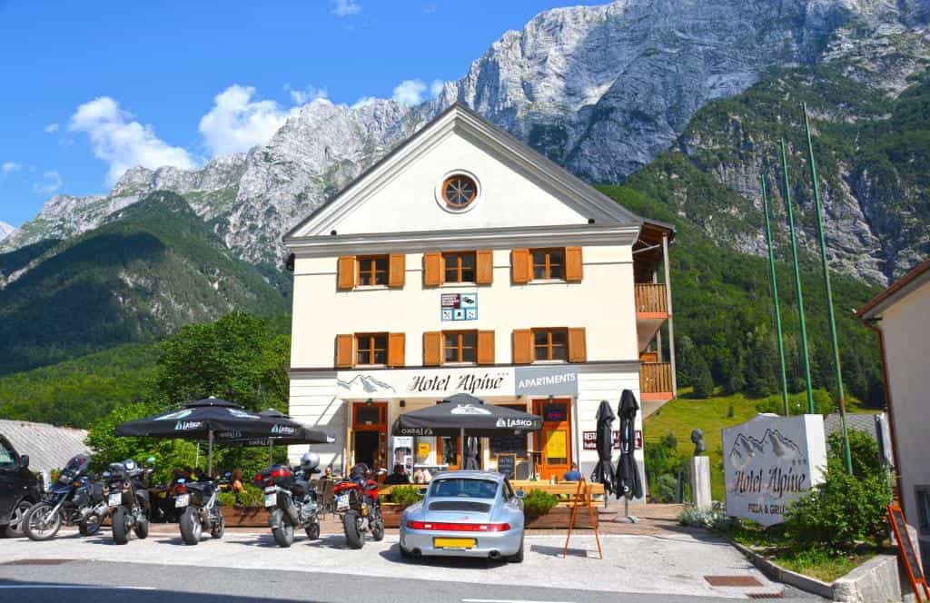 Hotel Alpine building with motorcycles and sports car parked against a backdrop of steep, forested mountains.