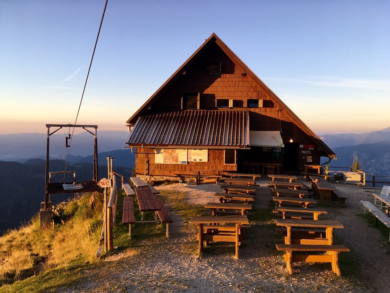Mountain hut with picnic tables at sunset overlooking layered mountain ranges, near Koča na Golici.