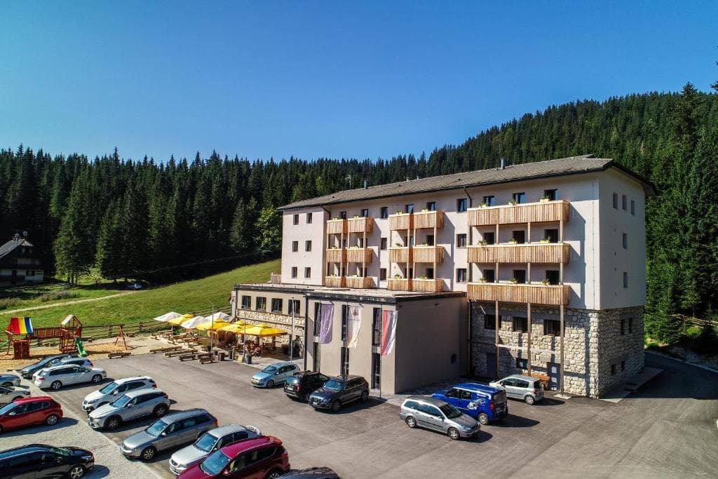 Hotel Pokljuka building with wooden balconies, parking lot, and dense pine forest backdrop.