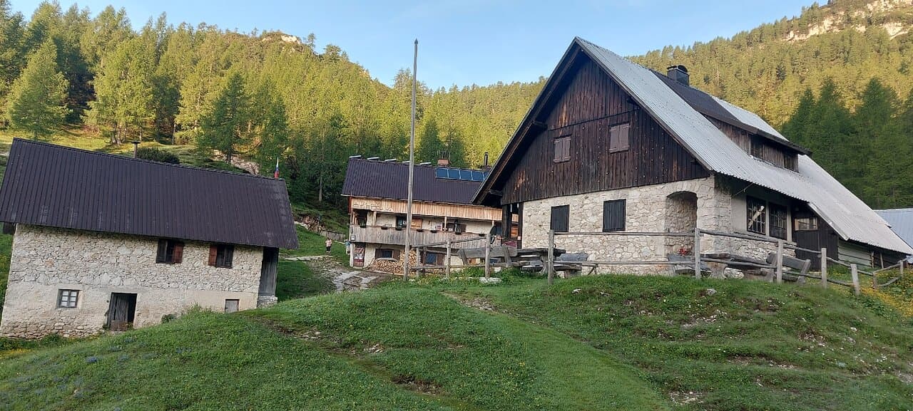 Stone mountain huts with wooden trim and metal roofs nestled on a grassy slope with pine forest background.