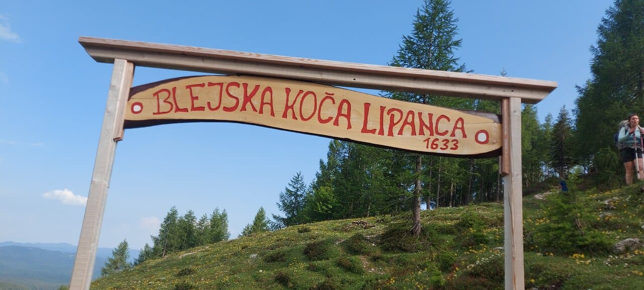Wooden sign for Blejska Koča Lipanca 1633 with hiker on grassy mountain slope