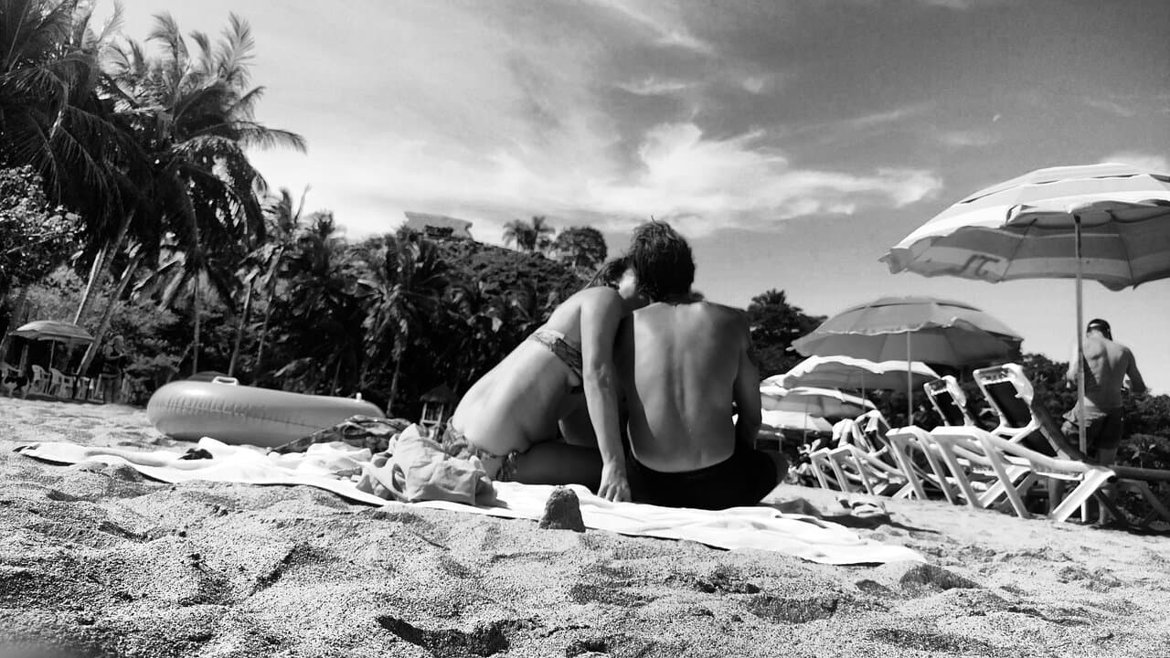 Couple relaxing on sandy beach with palm trees, beach chairs, and umbrellas in black and white.