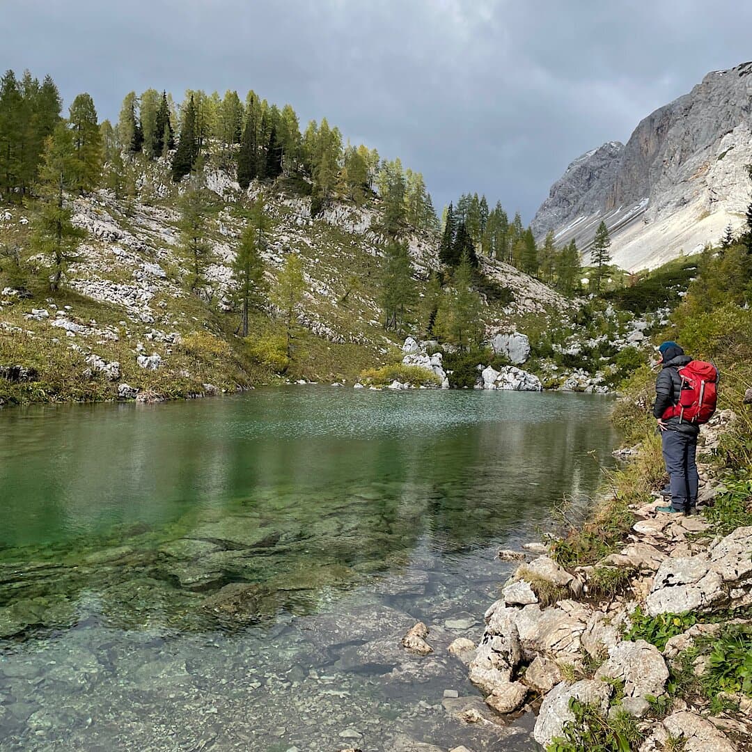 Hiker with red backpack viewing clear green water of an alpine lake surrounded by rocky slopes and trees.
