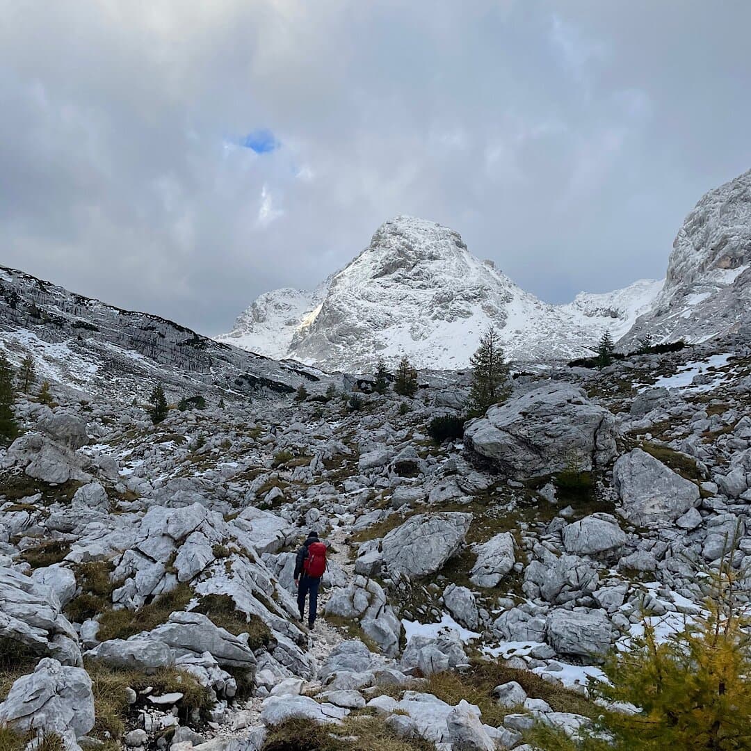Hiker with red backpack ascending rocky, snow-dusted terrain toward snow-capped mountains under cloudy sky.