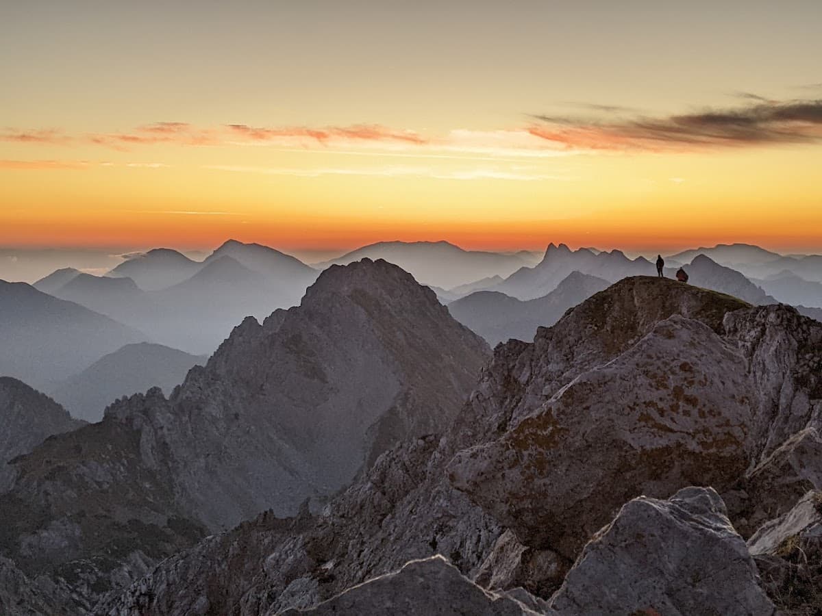 Hikers silhouetted on rocky mountain peak at sunrise over layered mountain ranges