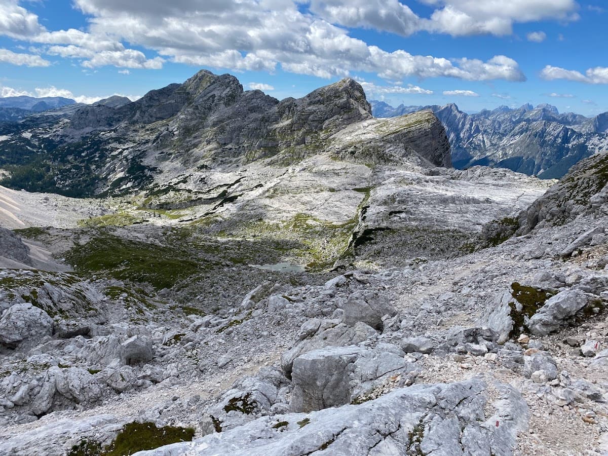Rocky alpine terrain with rugged peaks under a blue sky with white clouds, small tarn visible.