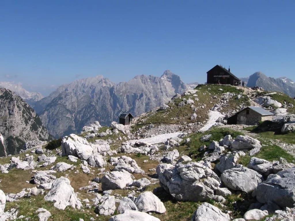 Zasavska Hut at Prehodavci on rocky alpine terrain with distant mountain range under clear blue sky.