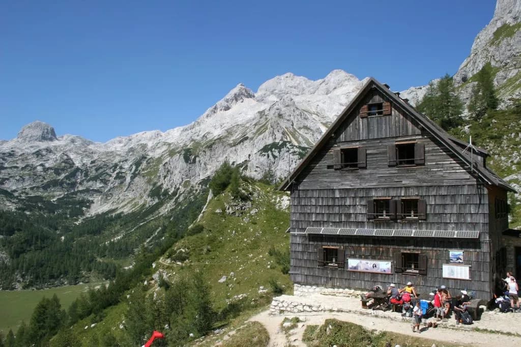 Vodnik lodge above Velo Polje with hikers resting against rugged limestone mountains.