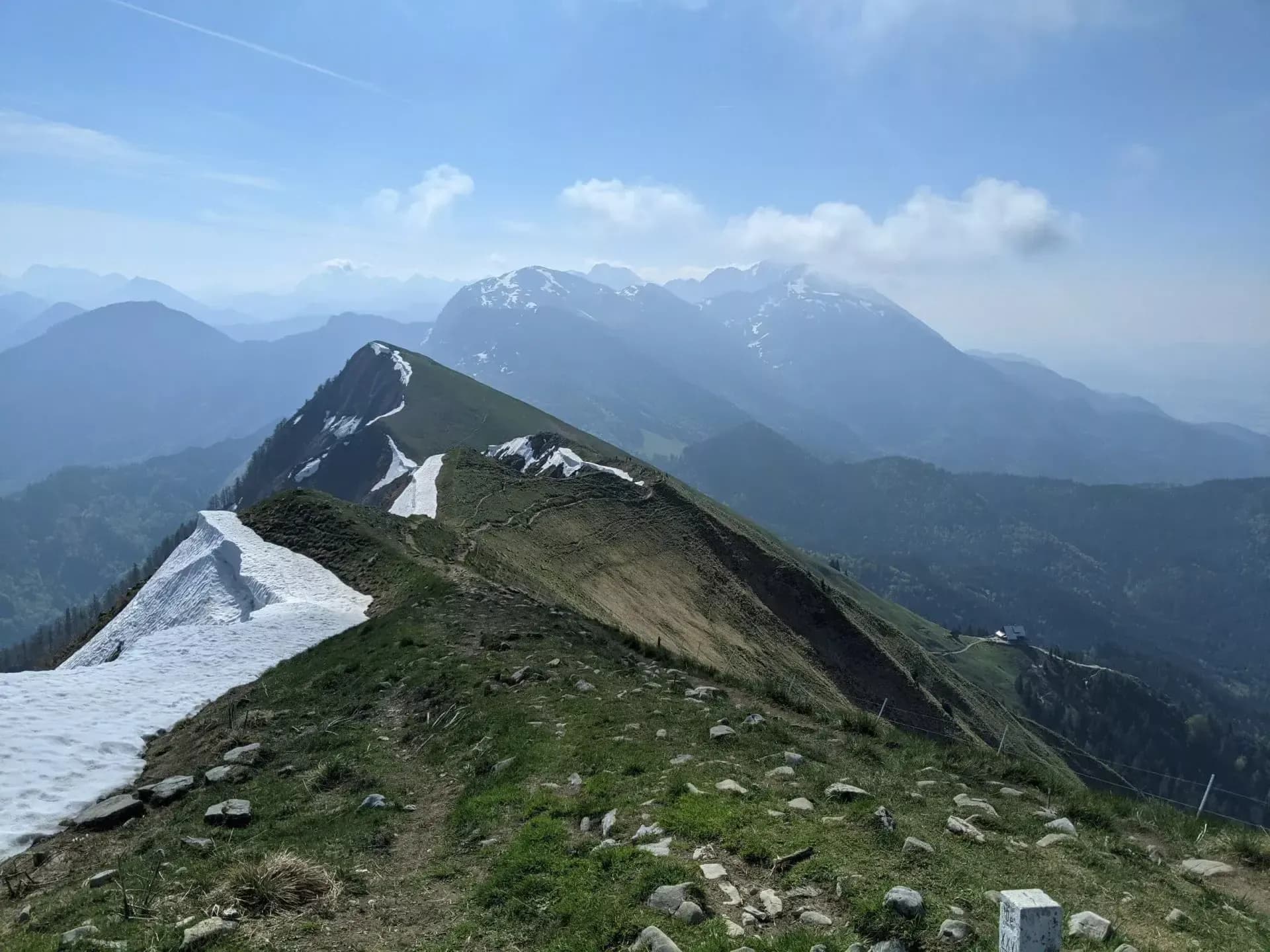 Hiking along a grassy ridge with snow patches towards hazy Karawanke mountains.