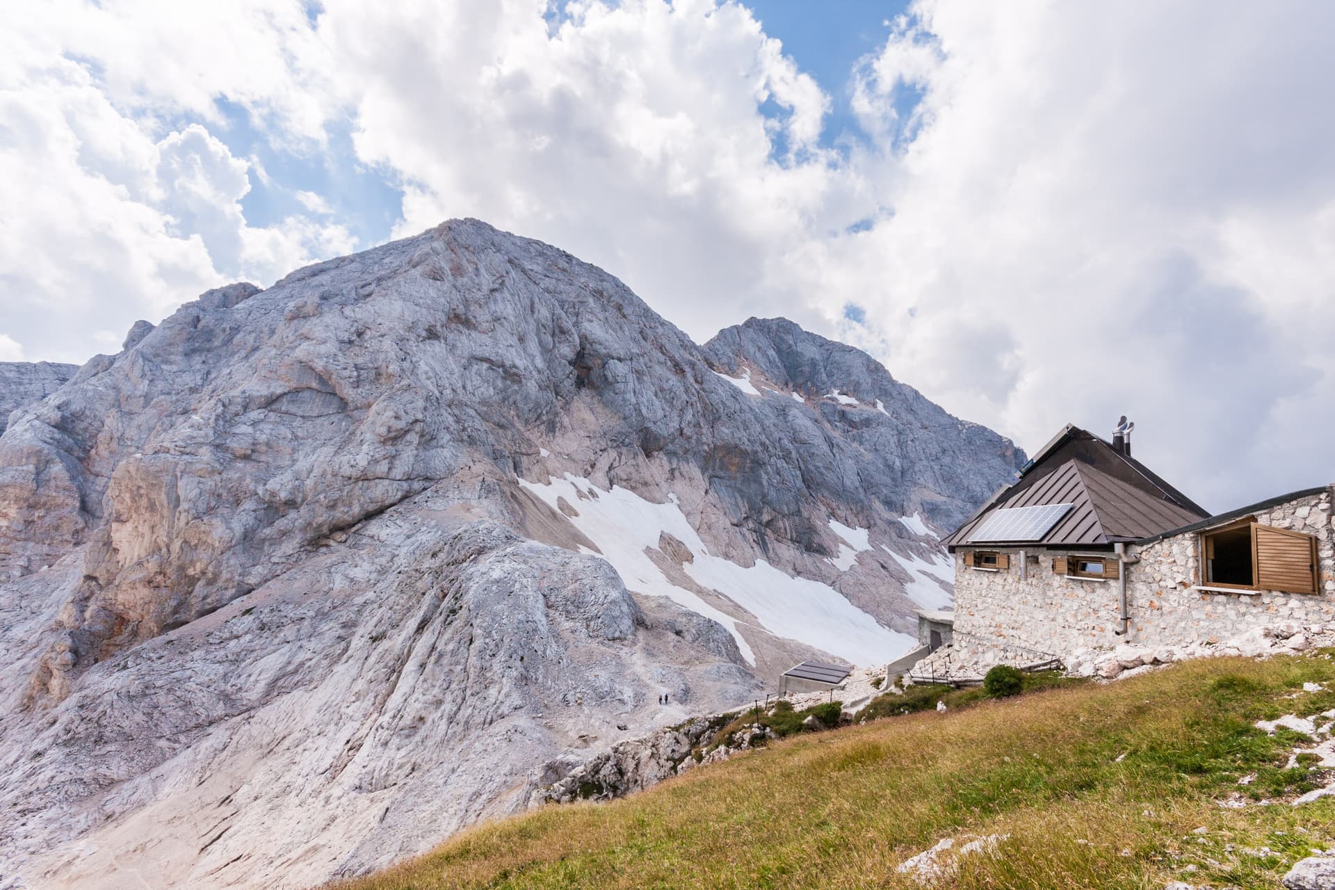 Mountain hut near massive rocky peaks with patches of snow under cloudy sky