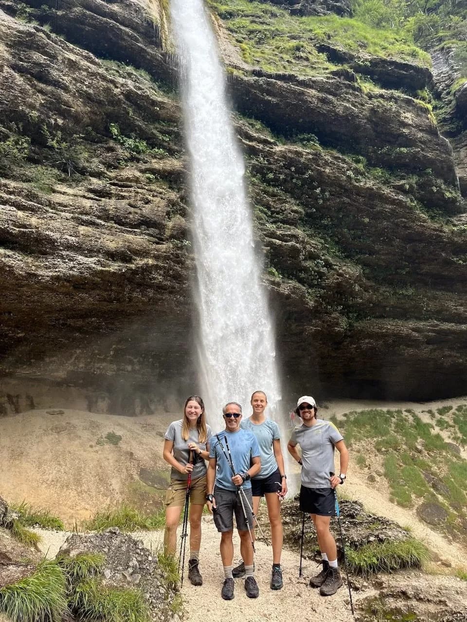 Hikers with trekking poles pose below Peričnik waterfall cascading down a rocky cliff face.