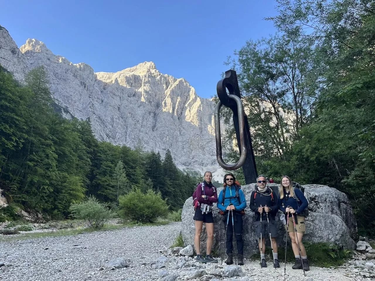 Hiking group with trekking poles stands by large carabiner sculpture near Vrata Valley mountains.
