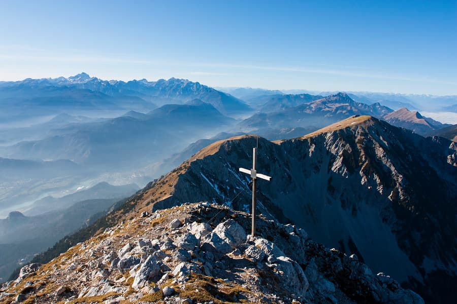 Summit cross on rocky mountain peak overlooking layered blue mountain ranges and valley haze.