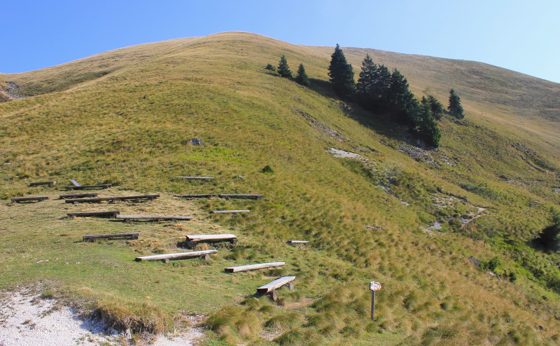 Wooden benches on grassy slope leading up to a rounded mountain under a clear blue sky.