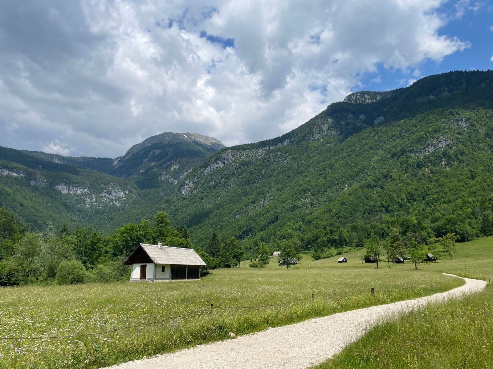 Small house in a green meadow with a gravel path and Julian Alps mountains in the background.