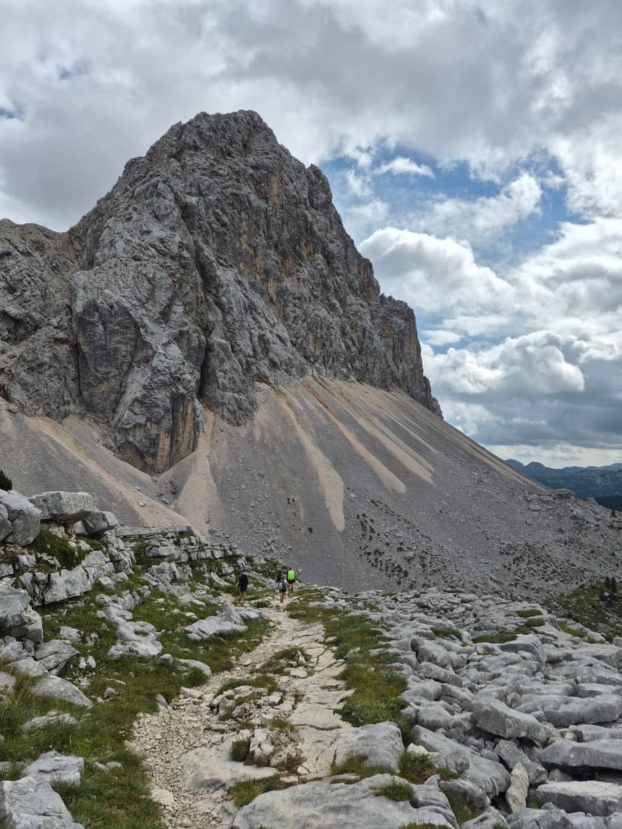 Hikers on a rocky trail beneath a massive mountain peak under a cloudy sky