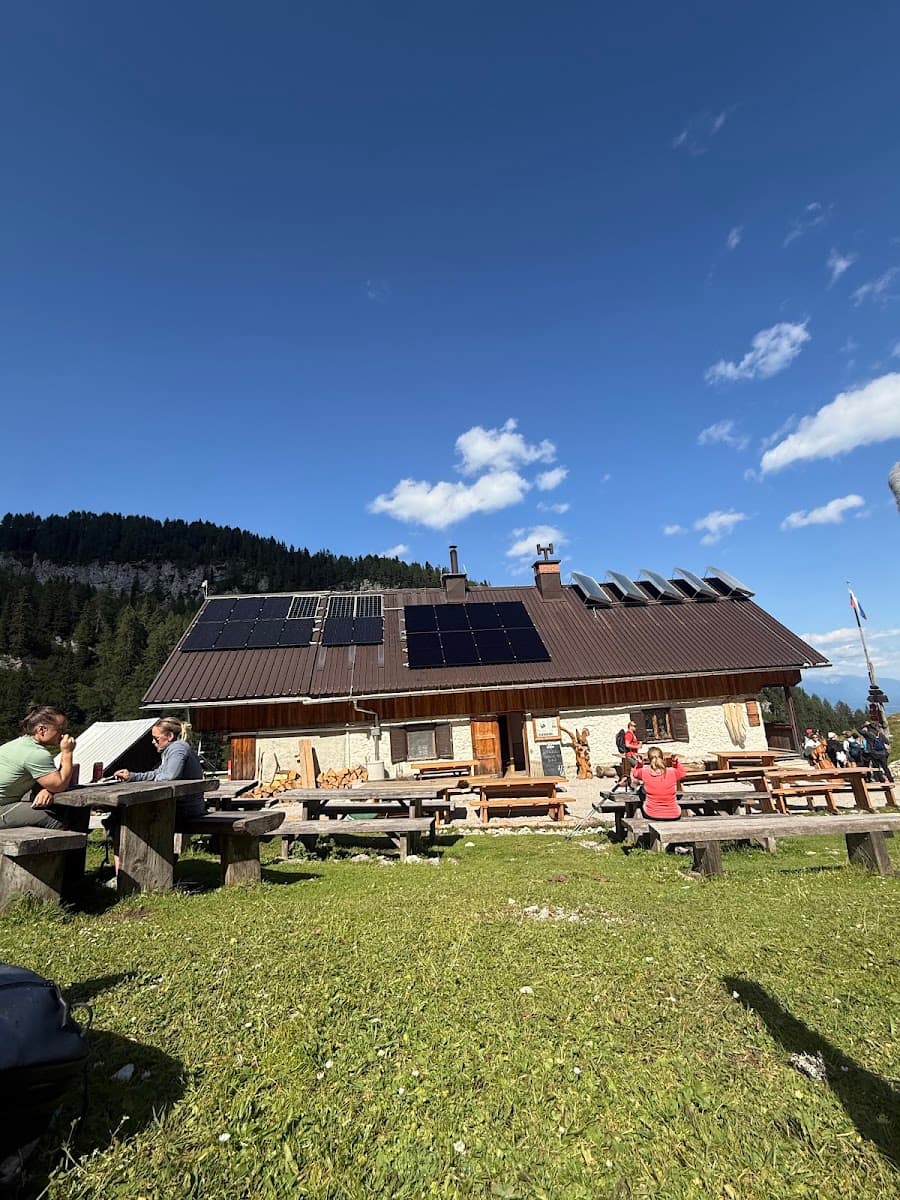 Alpine mountain hut with solar panels and outdoor picnic tables on a grassy slope under a blue sky