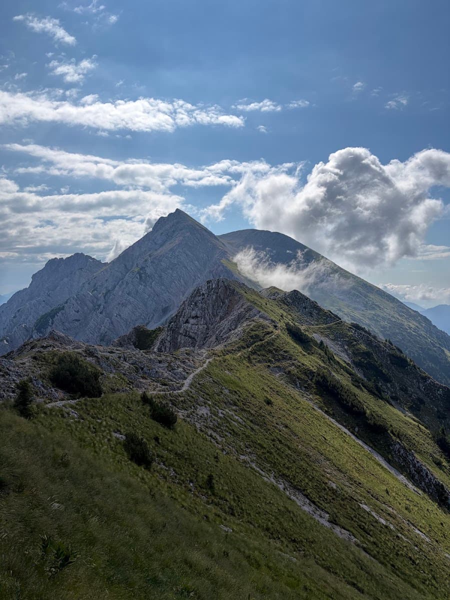 Alpine mountain peaks with hiking trail and green grassy slopes under a cloudy blue sky