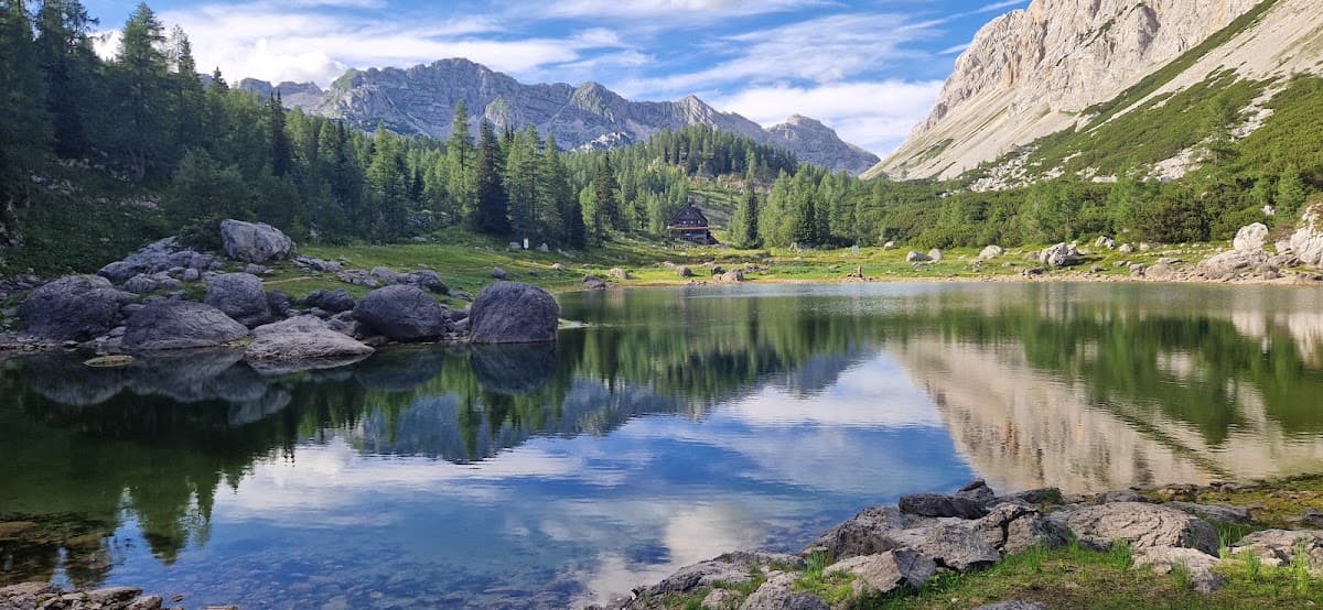 Alpine lake reflecting surrounding mountains, forest, and small wooden cabin with boulders in the foreground