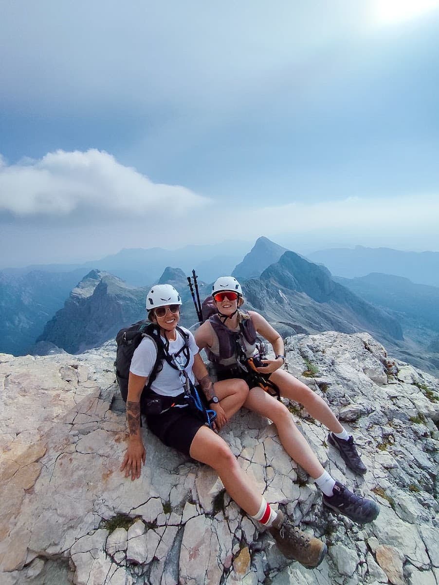 Two hikers wearing helmets and backpacks sitting on a rocky mountain summit with panoramic views