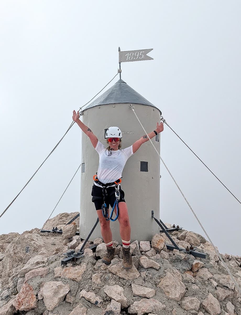 Climber celebrating on a rocky mountain summit beside a metal shelter with an 1895 flag