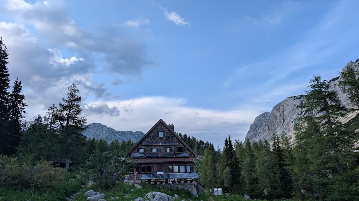 Wooden alpine cabin surrounded by pine trees and mountain peaks under a cloudy blue sky