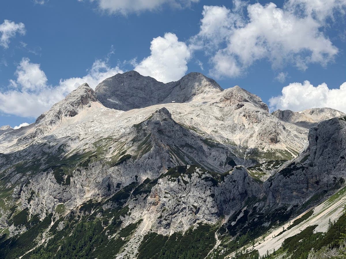 Rugged alpine mountain peaks with rocky slopes and scattered pine trees under a blue sky with white clouds