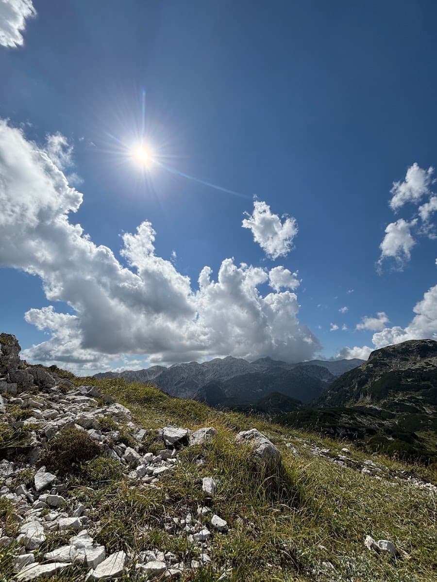 Mountain landscape with rocky slope, green grass, and bright sun shining through clouds in blue sky