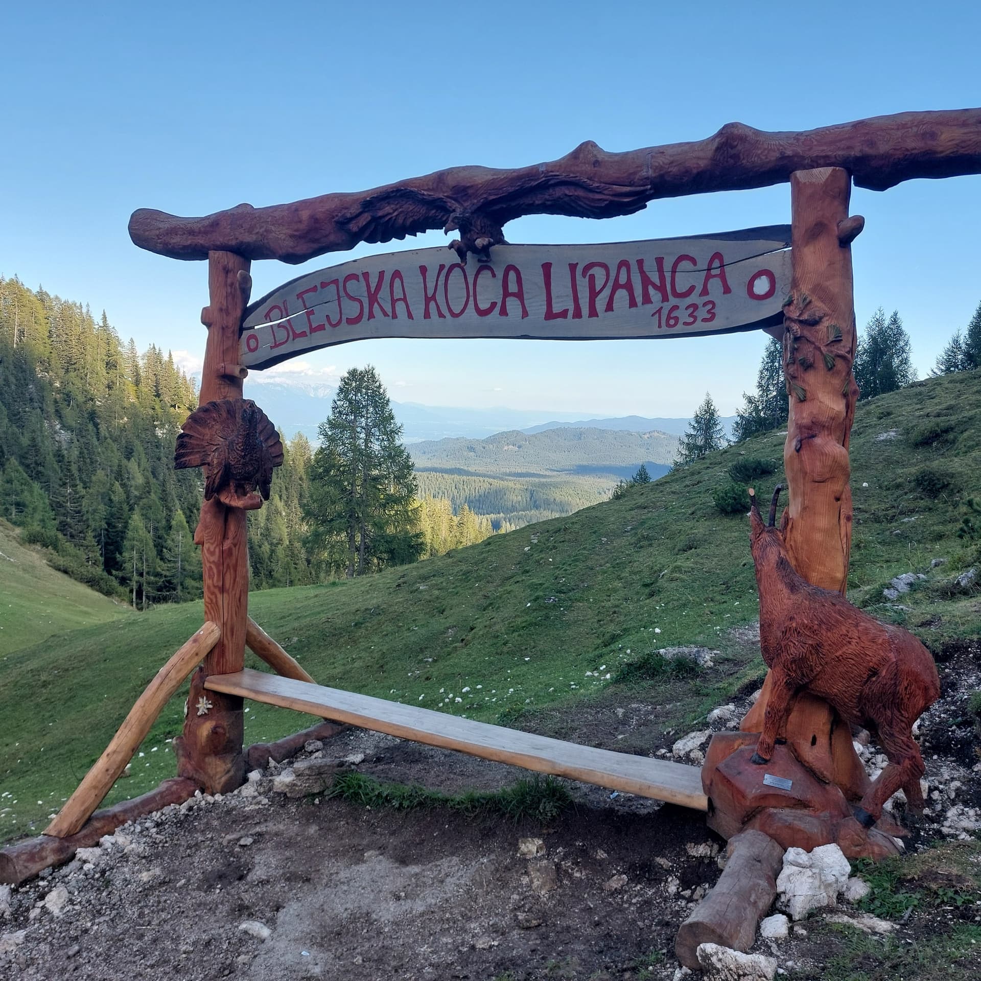 Carved wooden sign at Blejska Koca Lipanca mountain hut with alpine forest and mountain backdrop