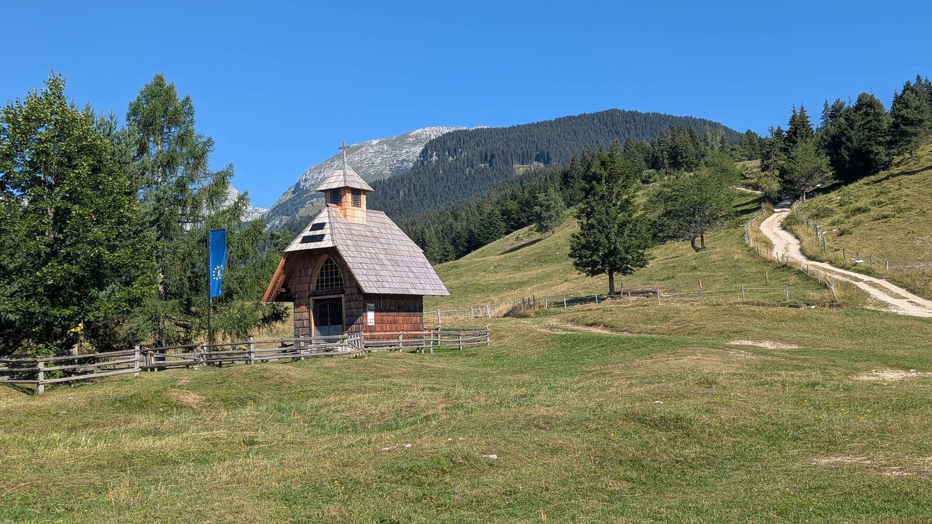 Wooden chapel on alpine mountain pasture with dirt path and forest