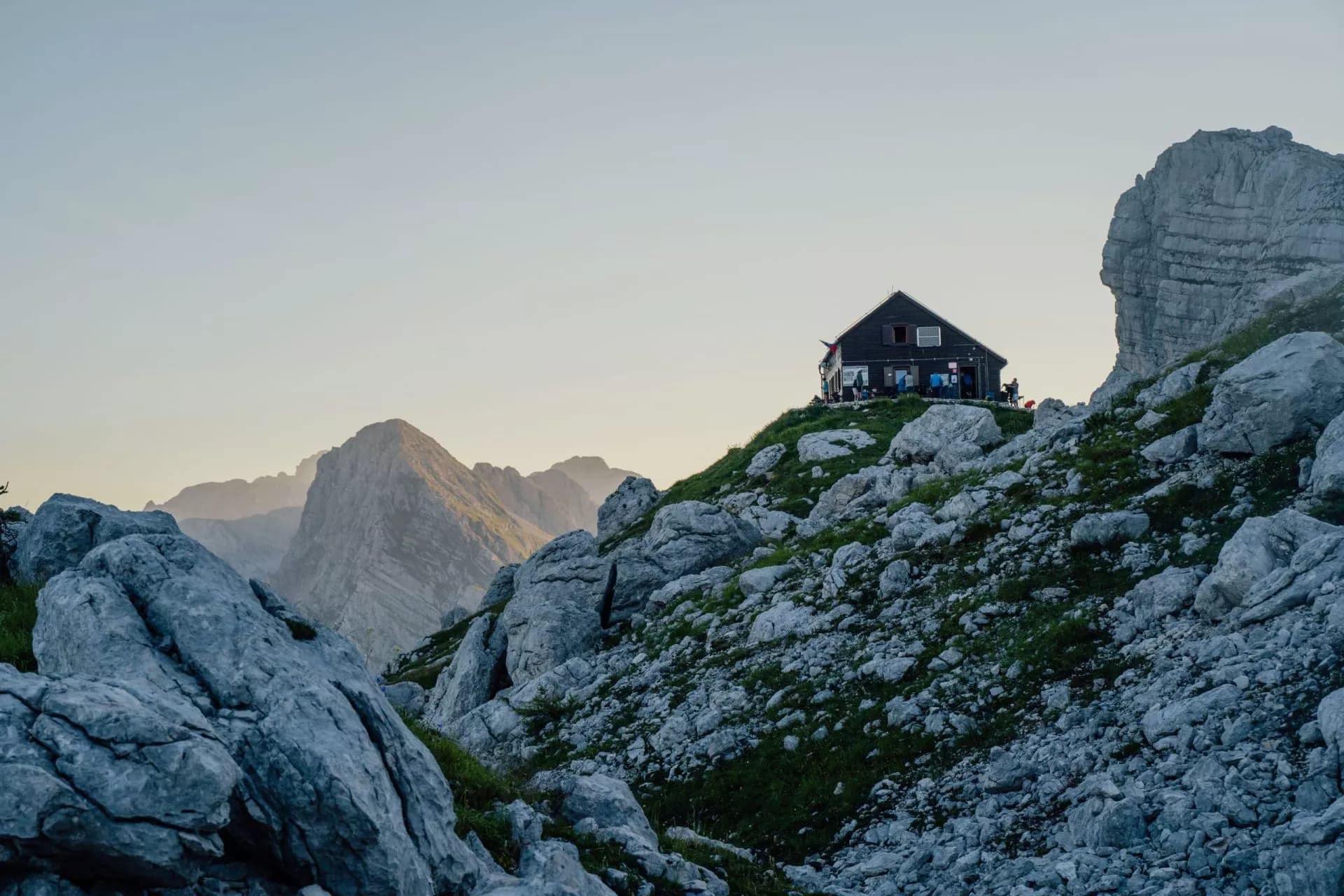 Mountain hut on rocky slope with hikers, set against hazy peaks at dawn or dusk