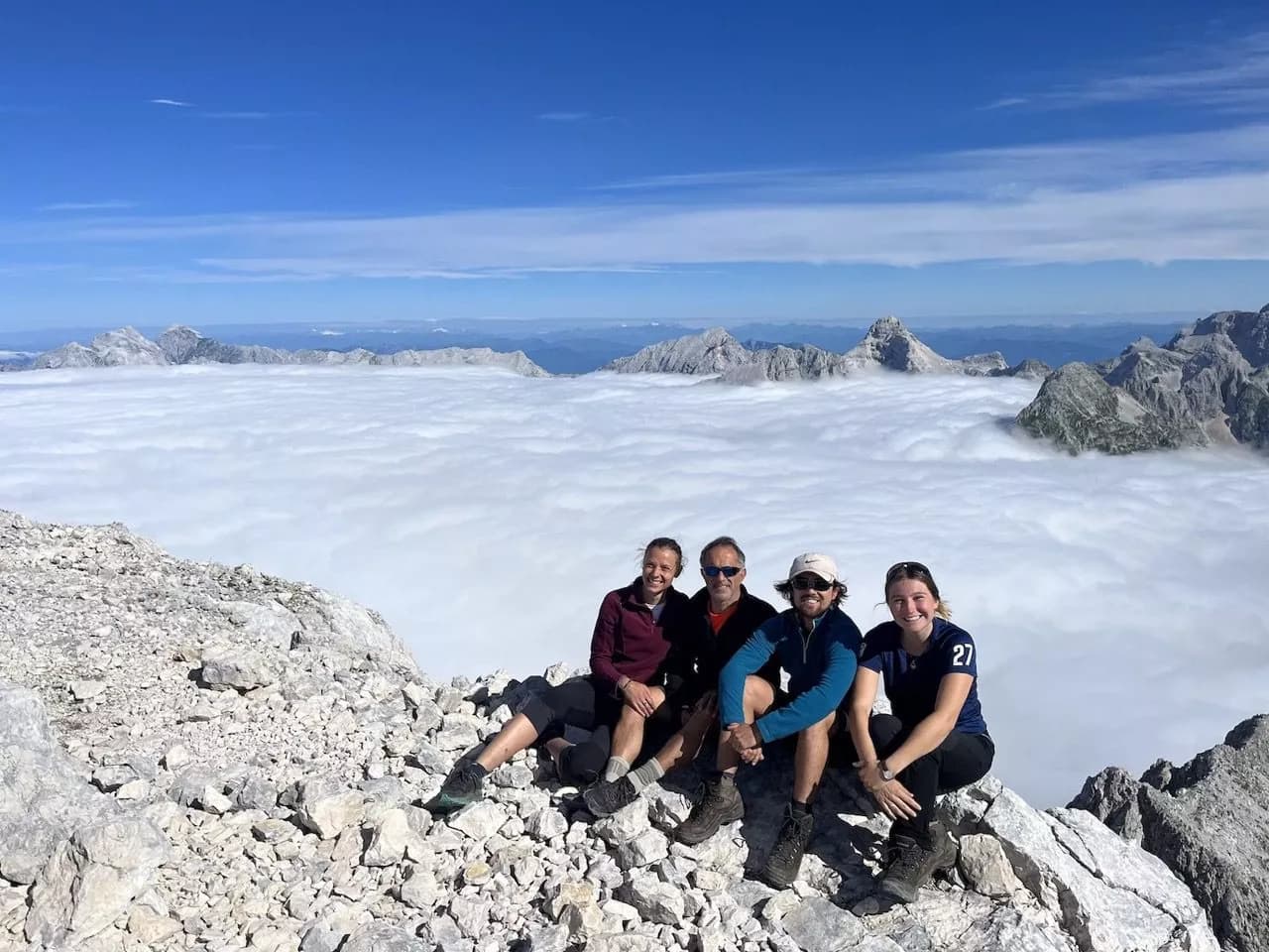 Hikers resting on rocky summit above a sea of fog with distant mountain peaks under blue sky.
