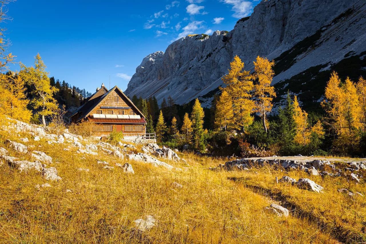 Triglav Lakes Lodge in autumn with golden trees against steep grey mountains under blue sky.