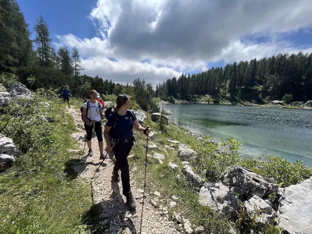 Hikers with trekking poles walk a rocky path beside a clear alpine lake surrounded by forest.