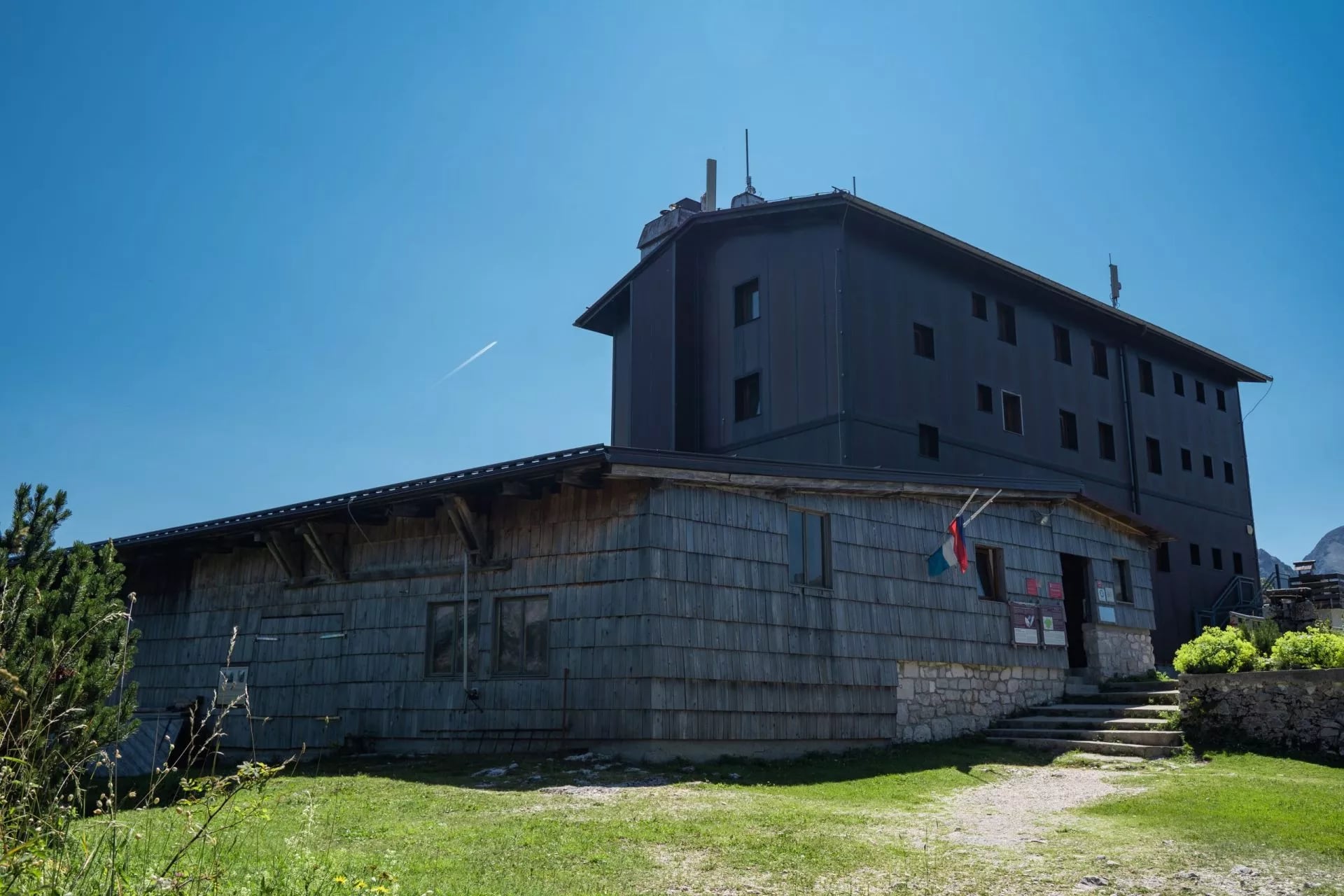 Mountain hut with wooden and dark siding under a clear blue sky with a contrail.