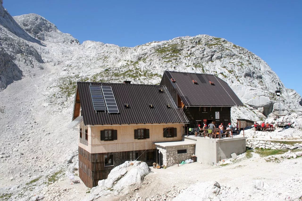 Mountain hut with solar panels nestled among rocky, high alpine terrain under a clear blue sky.
