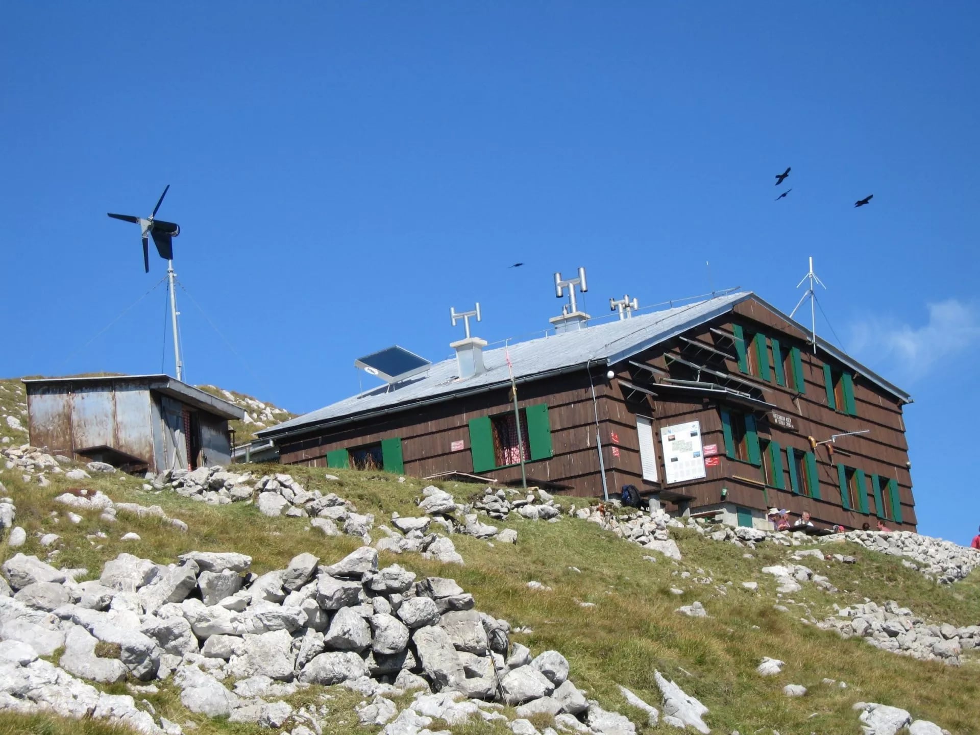 Preseren Hut on Stolu mountain with solar panels, wind turbine, and birds flying.