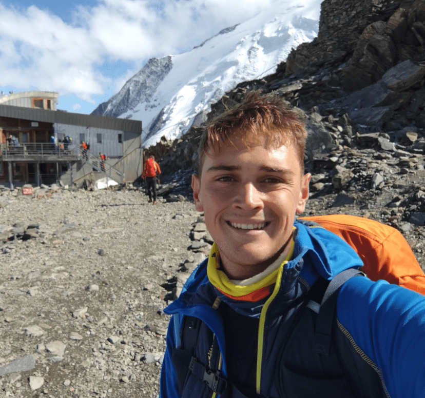 Hiker taking a selfie in front of a mountain hut with snow-capped peaks in the background.