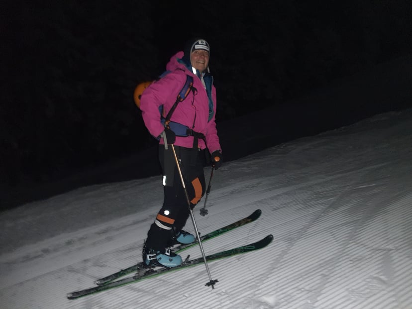 Skier in a pink jacket and backpack standing on a groomed snowy slope at night.