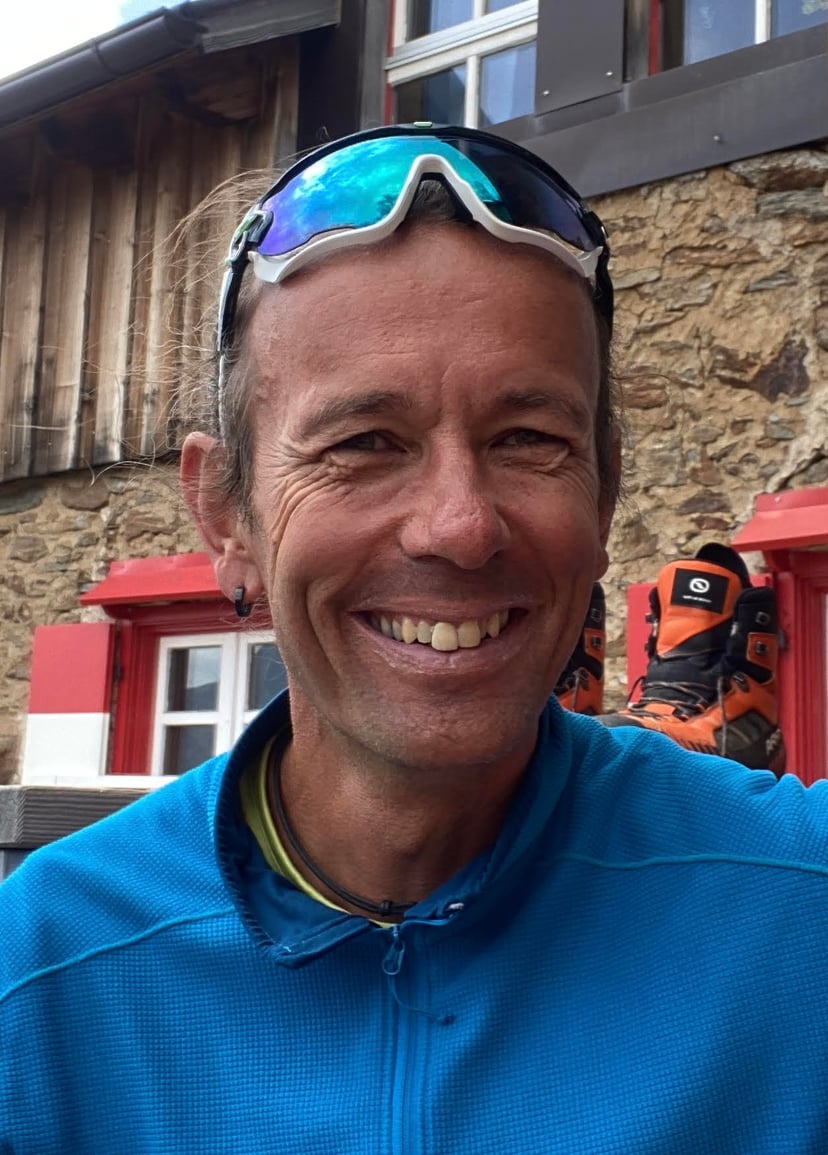 Smiling man wearing sports sunglasses and a blue fleece jacket in front of a stone building.