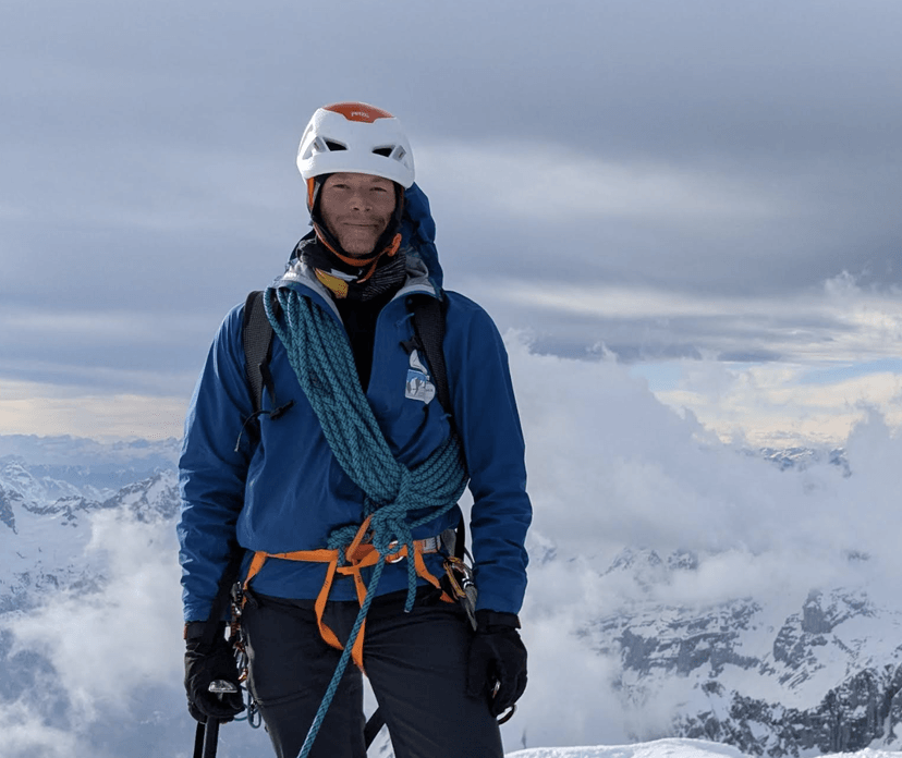 Mountaineer wearing a helmet and climbing gear standing on a snowy mountain peak above the clouds.