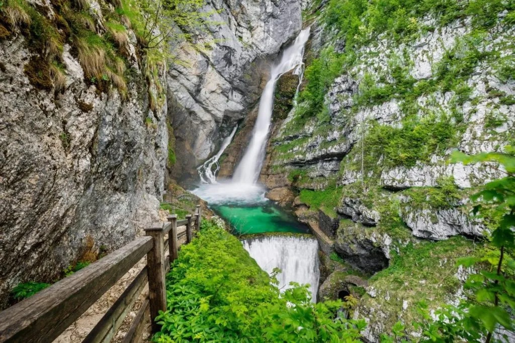 Savica Waterfall cascading into a turquoise pool with a wooden walkway along the cliffside.