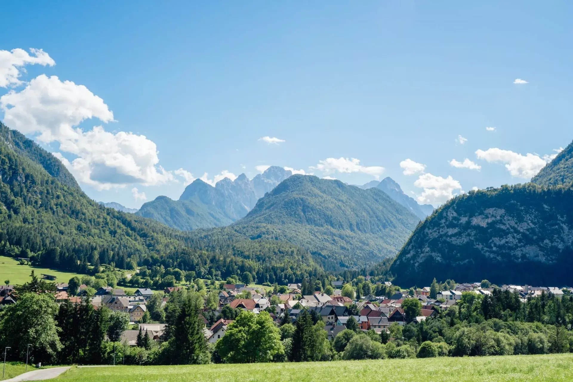 Village nestled in a green valley surrounded by forested mountains under a blue sky.