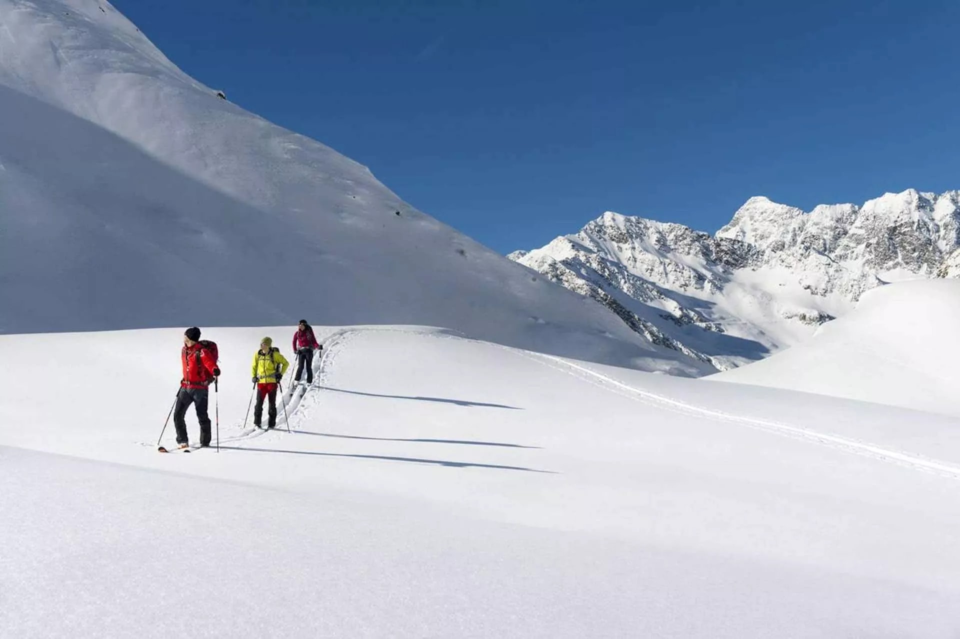 Ski touring group ascending snowy slope below jagged, snow-covered Julian Alps peaks under blue sky.