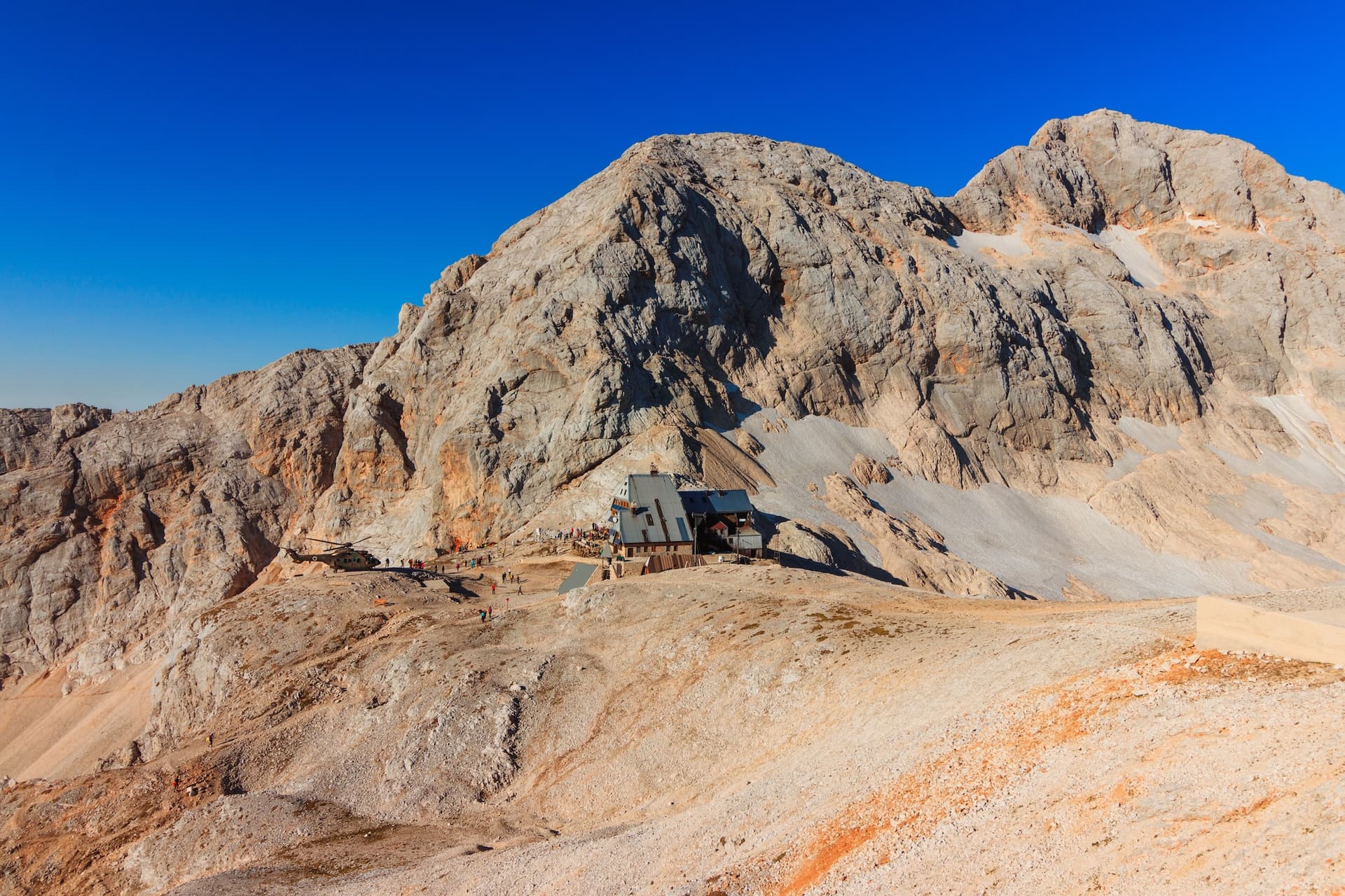 Mountain hut below Triglav peak with helicopter landing on rocky terrain under clear blue sky.