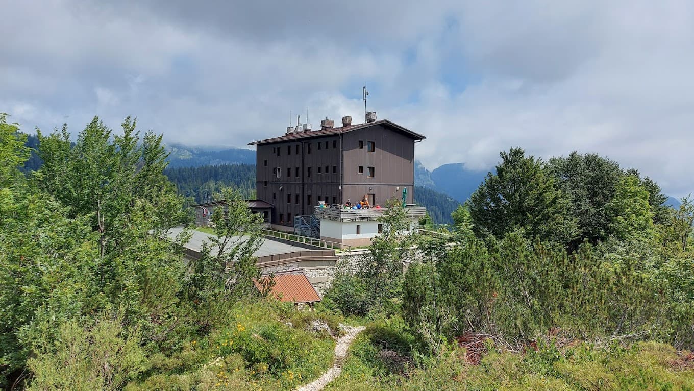 Mountain lodge surrounded by green trees with hikers on the terrace under cloudy skies.