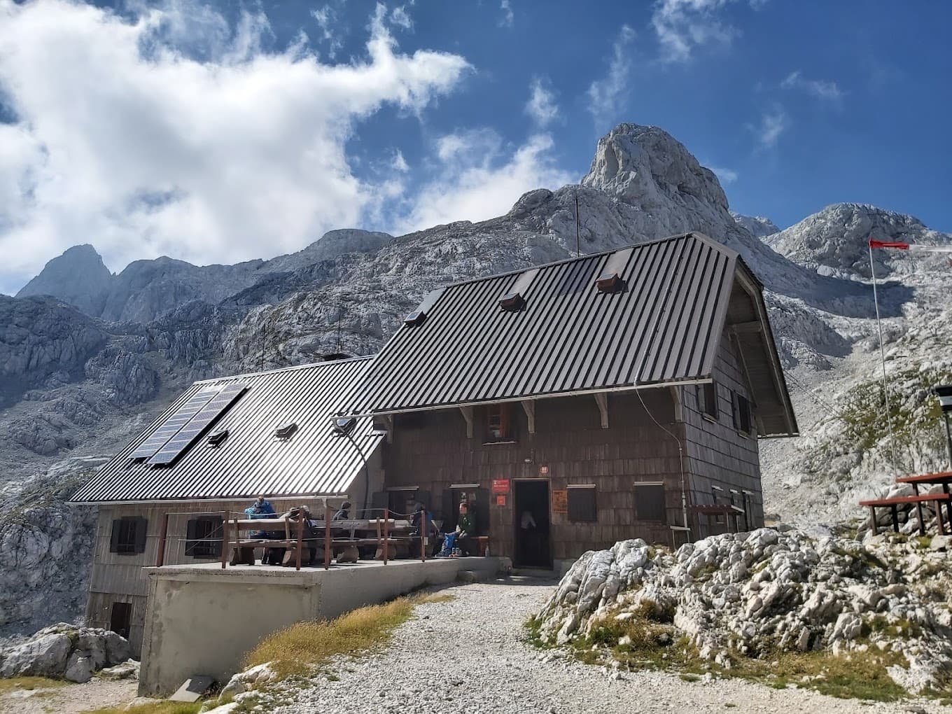 Mountain hut with solar panels on metal roof below jagged peaks, possibly Koča na Doliču.