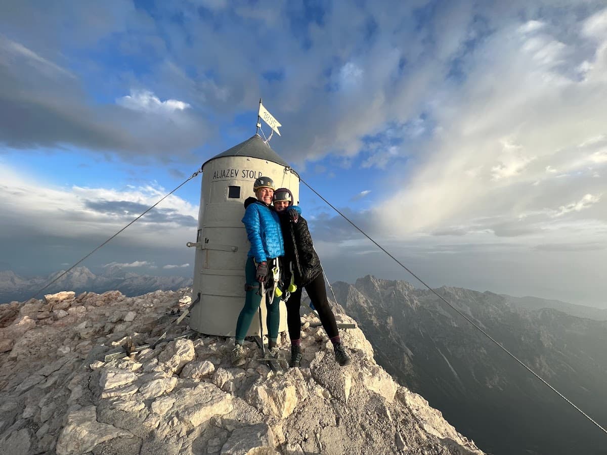 Two climbers pose by Aljažev Stolp on a rocky mountain summit under a cloudy sky.